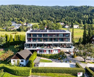 A modern building with several floors, surrounded by green landscape and trees. Guests are sitting on the terrace enjoying the view.