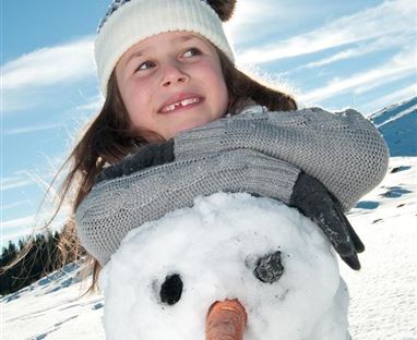 Ein Mädchen steht lächelnd neben einem Schneemann im Schnee. Der Schneemann hat eine Karottennase und Steinaugen.