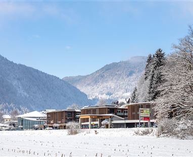 Eine verschneite Landschaft mit modernen Gebäuden im Vordergrund. Sanfte Hügel und schneebedeckte Bäume umgeben die Szenerie.