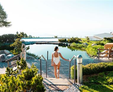 Un bel spazio esterno con un laghetto e tanto verde. Una donna in costume da bagno si dirige verso una terrazza in legno.