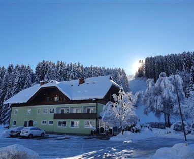 Ein schönes Gebäude im Schnee mit einer malerischen Waldlandschaft im Hintergrund. Die Sonne strahlt hinter den Bäumen hervor und schafft eine friedliche Winteratmosphäre.