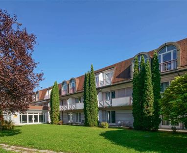 Un edificio moderno con più piani e grandi finestre. Circondato da alberi verdi e un prato curato.
