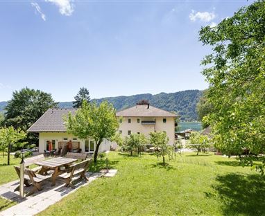 Ein schönes Haus mit einem großen Garten und einer Terrasse. Im Hintergrund sind Berge und ein klarer Himmel zu sehen.