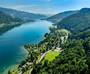 Ein wunderschöner See umgeben von grünen Bergen und Wäldern. Im Vordergrund sind einige Häuser und ein gepflasterter Weg zu sehen.