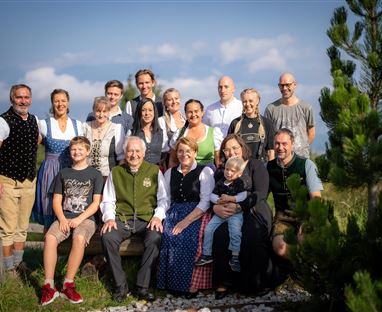 Eine Gruppe von Menschen in traditioneller Kleidung steht draußen in der Natur. Im Hintergrund sind Bäume und ein klarer Himmel zu sehen.