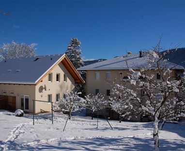 Zwei Häuser im Schnee unter einem blauen Himmel. Der Garten ist mit Schneedecke bedeckt und es gibt einige schneebedeckte Bäume.