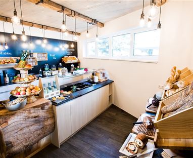 Ein gemütlicher Verkaufsraum mit einer Auswahl an frischen Lebensmitteln. An einem Holzstand gibt es Obst, Backwaren und Getränke.