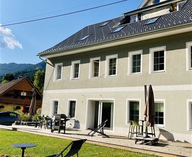 Ein modernes Gebäude mit einem großen Garten und Terrassenmöbeln. Der Himmel ist klar und es gibt einige Berge im Hintergrund.