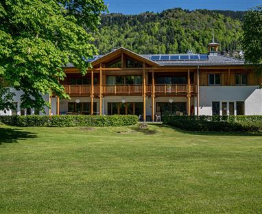 Ein modernes Gebäude mit Holzverkleidung und großzügigem Balkon. Umgeben von grünem Rasen und Bäumen in einer bergigen Landschaft.