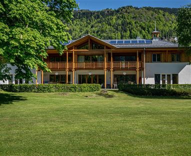 Ein modernes Gebäude mit Holzverkleidung und großzügigem Balkon. Umgeben von grünem Rasen und Bäumen in einer bergigen Landschaft.