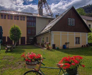 Ein rustikales Fahrrad mit blühenden roten Geranien steht auf einer Wiese. Im Hintergrund sind alte Gebäude und ein Förderturm zu sehen.