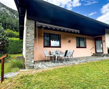 Ein modernes Haus mit einer Terrasse und Garten. Im Hintergrund sind grüne Hügel und blauer Himmel zu sehen.