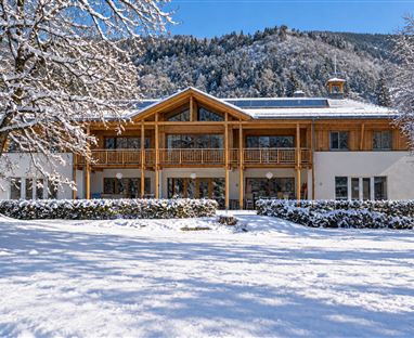 A modern wooden house in the snow, surrounded by snow-covered trees. In the background, gentle hills and a blue sky can be seen.