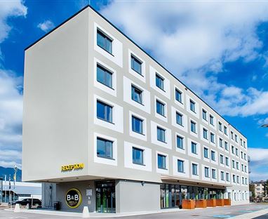 Ein modernes Hotelgebäude mit fünf Etagen und großen Fenstern. Die Fassade ist hell und schlicht, umgeben von einem klaren Himmel.