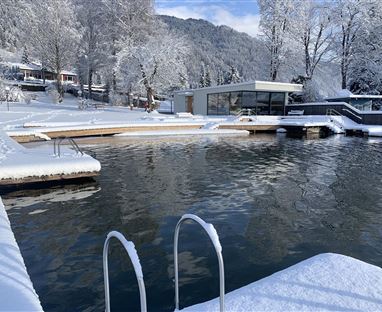 Un bel lago invernale con una riva coperta di neve e un edificio moderno sullo sfondo. Il paesaggio è tranquillo e gli alberi sono coperti di neve.