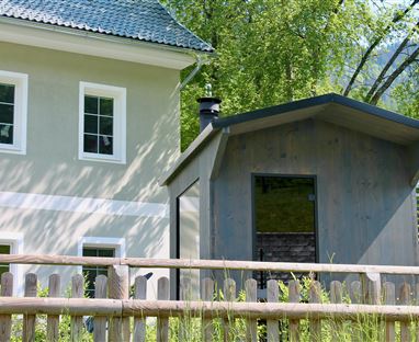 Ein modernes Haus in hellen Farben steht neben einem kleinen Schuppen. Die Umgebung ist von Grünpflanzen und einem Zaun umgeben.