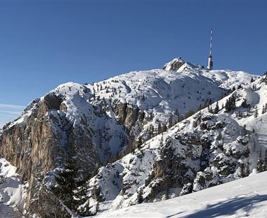 Una montagna coperta di neve con un traliccio sulla cima. Il cielo blu chiaro offre un bel contrasto con il paesaggio invernale.