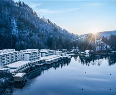 Un paesaggio invernale tranquillo con un hotel sulle rive di un lago calmo. Il sole sorge dietro le montagne e tinge la scena di una luce morbida.