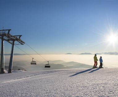 Un paesaggio montano innevato con una funivia e due sciatori. Il cielo è sereno e il sole splende radioso.