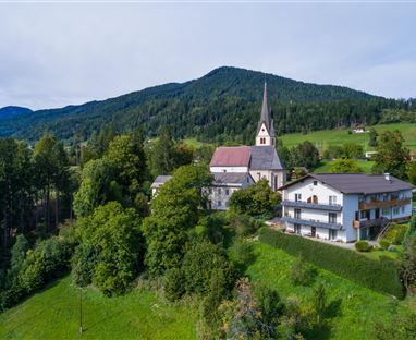 Eine malerische Landschaft mit einer Kirche und einem ehemaligen Bauernhaus. Im Hintergrund erstrecken sich grüne Hügel und Wälder.