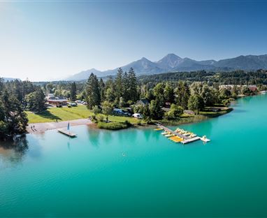 Ein ruhiger See mit klarem, türkisfarbenem Wasser und grünen Uferbereichen. Im Hintergrund sind beeindruckende Berge und eine schöne Landschaft sichtbar.