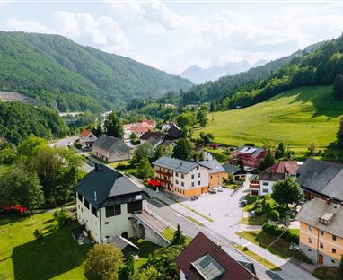 Ein malerisches Dorf in einer grünen Landschaft mit Blick auf die Berge. Die Häuser sind umgeben von Wiesen und Bäumen und strahlen eine ruhige Atmosphäre aus.