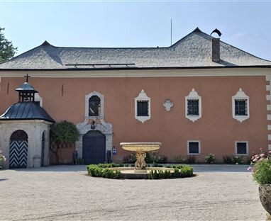 A historical building with a red facade and a lovely garden. In the foreground, there is a fountain surrounded by flower beds.