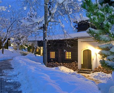 A winter village with snow-covered trees and a cozy wooden house. Soft light shines from the windows, illuminating the snowy path.