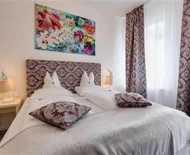 Ein stilvolles Schlafzimmer mit einem großen Bett und dekorativen Kissen. Die Wände sind mit ansprechenden Vorhängen und einem bunten Wandbild geschmückt.