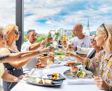 Un gruppo di persone alza i bicchieri e festeggia durante un pasto all'aperto. Sullo sfondo ci sono volti sorridenti e una vista accattivante.