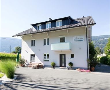 Ein modernes Haus mit einem Balkon und einer einladenden Eingangstür. Umgeben von blühenden Pflanzen und einem klaren Himmel.