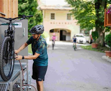 Ein Mann repariert ein Fahrrad in einer malerischen Umgebung. Im Hintergrund fahren weitere Radfahrer und es gibt grüne Bäume sowie Blumen.