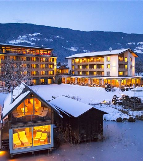 Un resort invernale con edifici illuminati nella neve. Sullo sfondo ci sono montagne e un lago tranquillo.