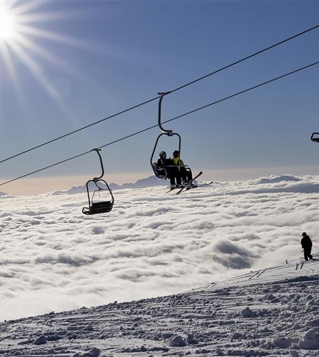 Un magnifico paesaggio montano con impianti di risalita che attraversano uno strato di nuvole. Il sole splende chiaro nel cielo.