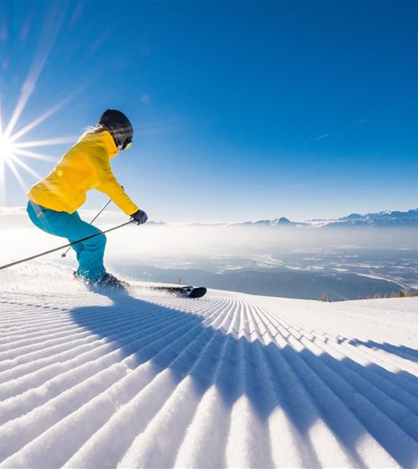 A skier in a yellow jacket is skiing on a freshly groomed slope. In the background, snow-covered mountains and a clear blue sky can be seen.