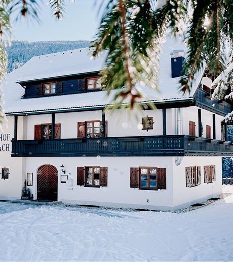 A charming guesthouse in winter, surrounded by snow. The facade is white with dark window frames and a traditional balcony.
