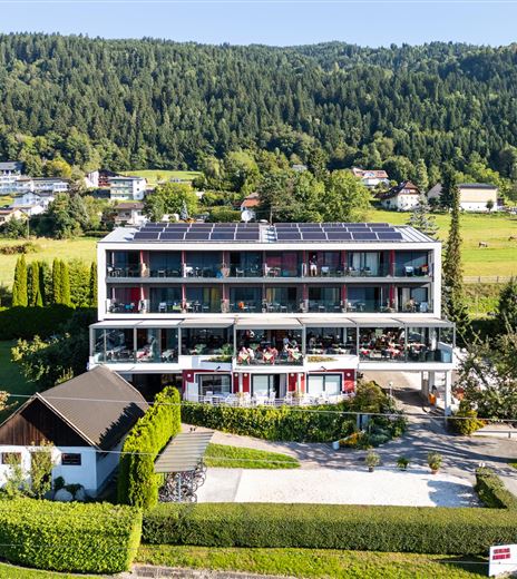 Ein modernes Gebäude mit mehreren Etagen, umgeben von grüner Landschaft und Bäumen. Auf der Terrasse sitzen Gäste und genießen die Aussicht.