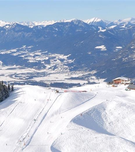 Eine schneebedeckte Berglandschaft mit mehreren Skipisten. Im Hintergrund sind majestätische Berge und ein klarer blauer Himmel zu sehen.