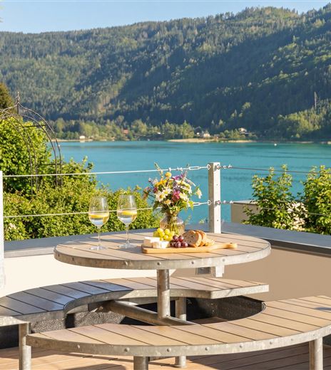 Eine schöne Terrasse mit einem runden Tisch, auf dem Wein und Obst stehen. Im Hintergrund liegt ein klarer See und grüne Berge.