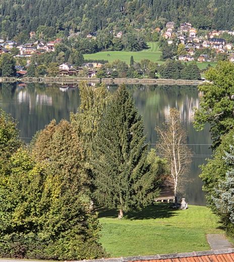 Ein ruhiger See umgeben von Bäumen und einer grünen Wiese. Im Hintergrund sind sanfte Hügel und Häuser zu sehen.