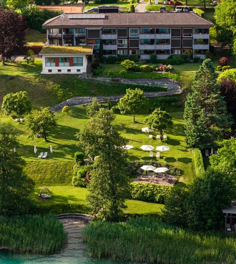 Eine grüne Gartenlandschaft mit Bäumen und Liegestühlen. Im Hintergrund befindet sich ein modernes Gebäude.