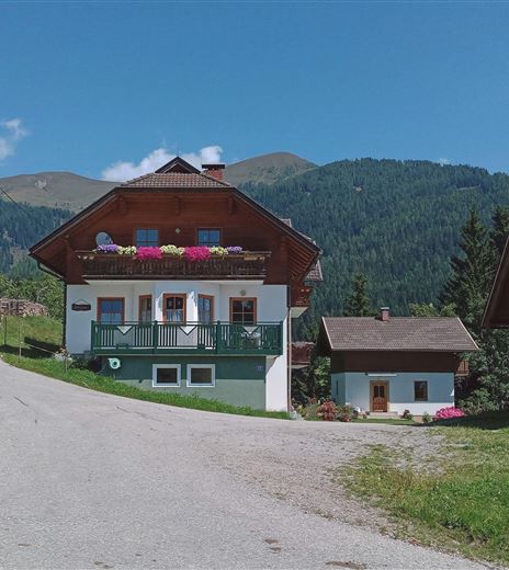 Ein charmantes Bauernhaus mit Balkonen und bunten Blumen. Im Hintergrund sind grüne Berge und ein klarer Himmel zu sehen.
