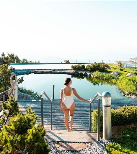 A beautiful outdoor area with a pond and lots of greenery. A woman in a swimsuit is walking towards a wooden terrace.