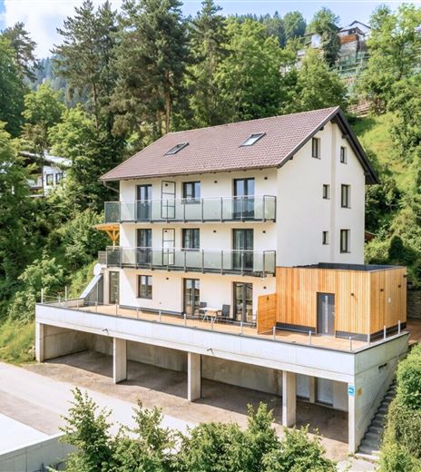 Ein modernes Haus am Hang, umgeben von Bäumen. Der äußere Bereich hat einen Balkon und eine Terrasse mit Platz zum Entspannen.