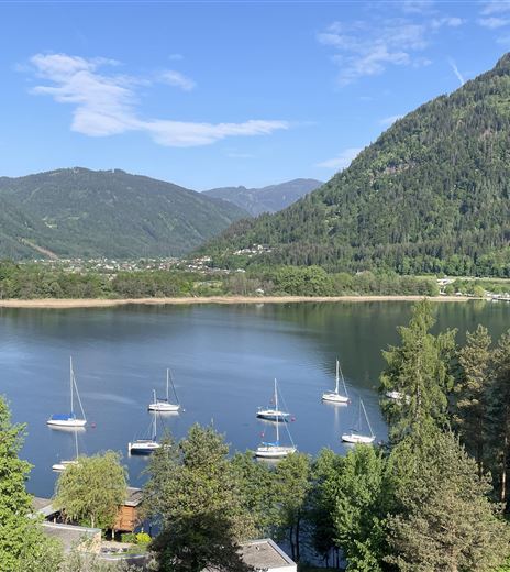 Ein klarer See umgeben von grünen Bergen und Bäumen. Segelboote liegen friedlich im Wasser.