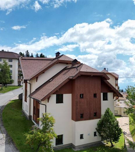 Una casa moderna con verniciatura in legno e bianco in un ambiente pittoresco. Sullo sfondo si possono vedere altri edifici e un paesaggio boschivo.