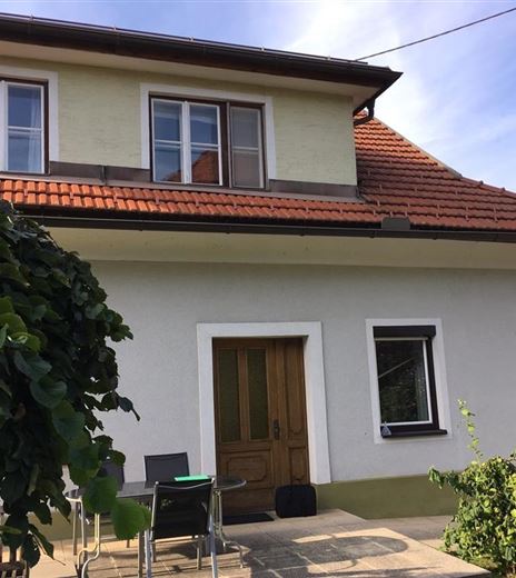 Ein gemütliches Haus mit einem roten Ziegeldach und einem kleinen Sitzbereich im Freien. Umgeben von grünen Pflanzen und einem klaren Himmel.