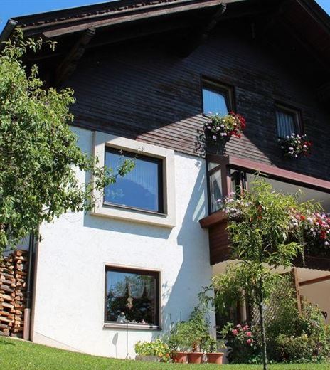 Ein gemütliches Haus mit einem Balkon voller Blumen. Der Garten ist grün und gut gepflegt, unter einem klaren blauen Himmel.