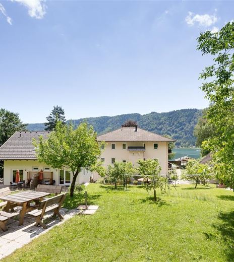 Ein schönes Haus mit einem großen Garten und einer Terrasse. Im Hintergrund sind Berge und ein klarer Himmel zu sehen.
