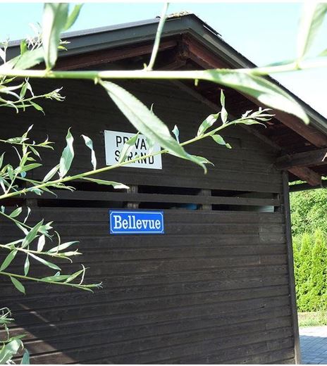Un edificio in legno con un'insegna che mostra "Bellevue". Circondato da piante verdi e un paesaggio curato.